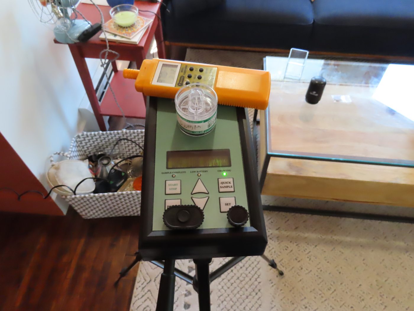 A spore-trap air sampling pump mounted on a tripod in a living room, a sample cassette fitted on top, with a sofa and side tables behind it