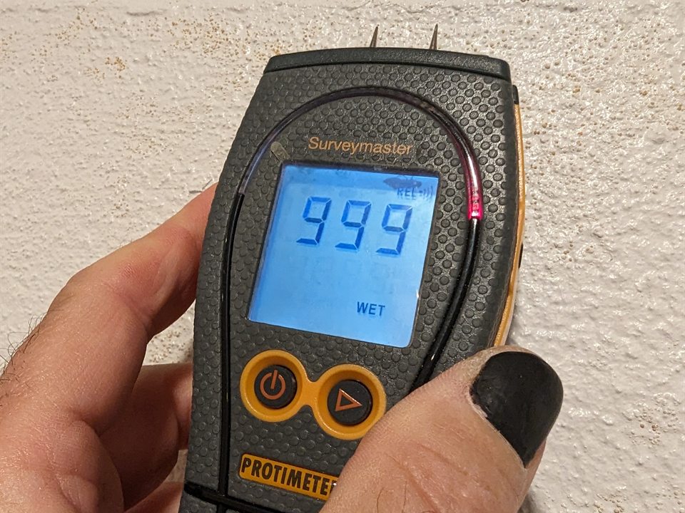 A moisture meter held against a painted masonry wall, its display reading 999 and WET
